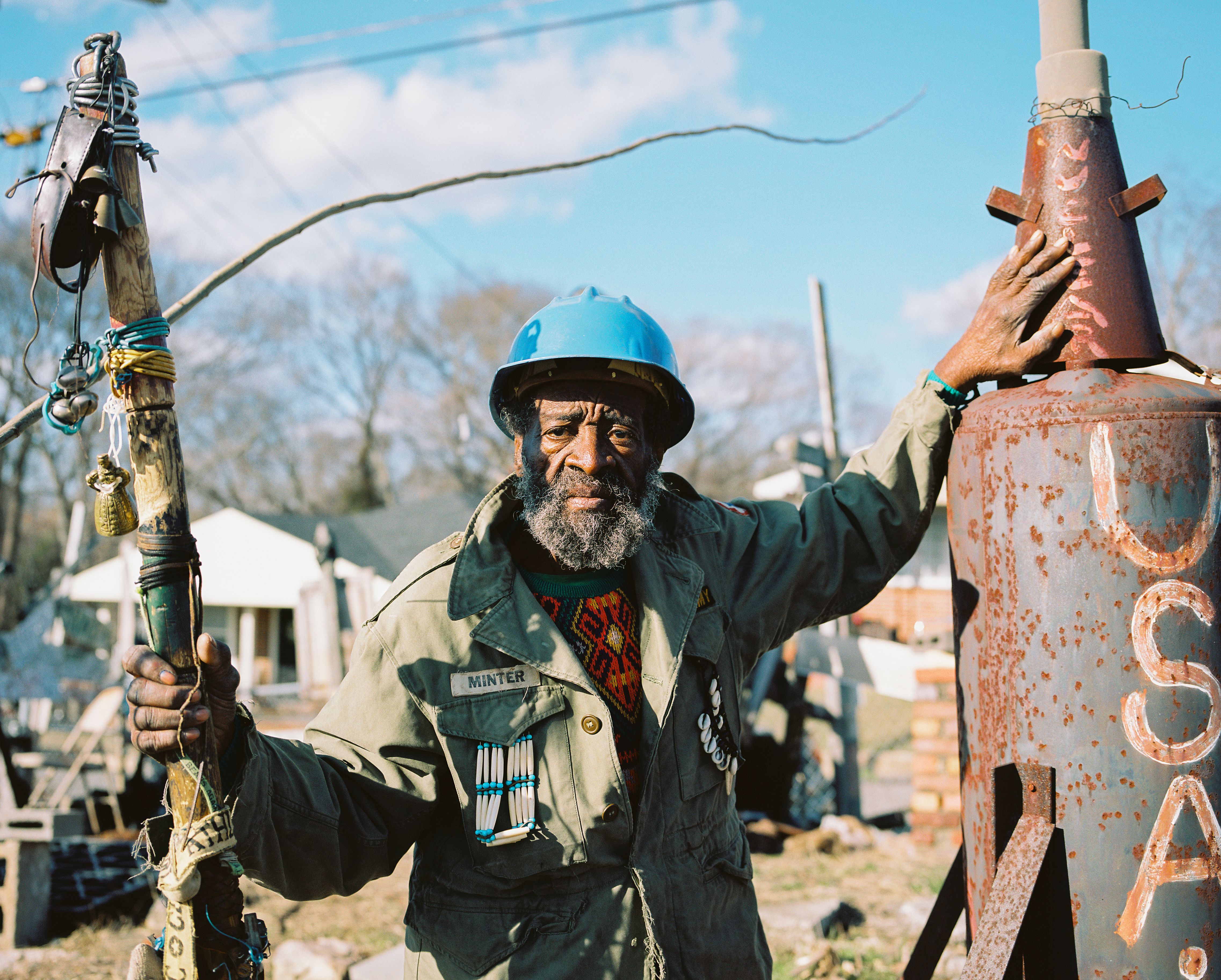 Joe holding a handmade sculptural staff, wearing a blue hardhat and old army jacket