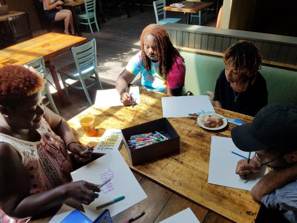 4 people gathered around a table with paper and drawing materials between them, in bright sunlight.