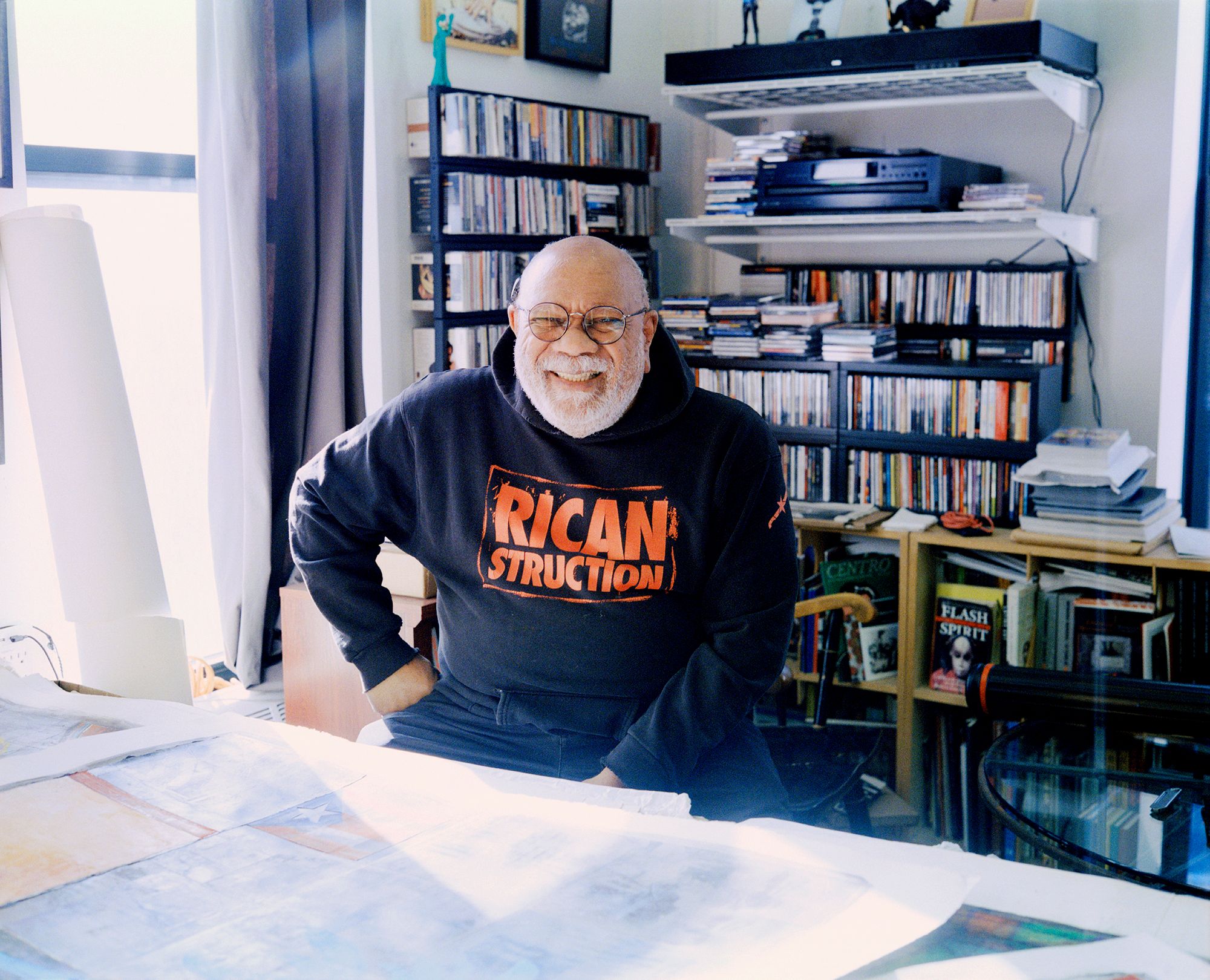 Juan Sánchez sitting inside his studio, smiling. wearing a sweatshirt which reads "RICAN STRUCTION"