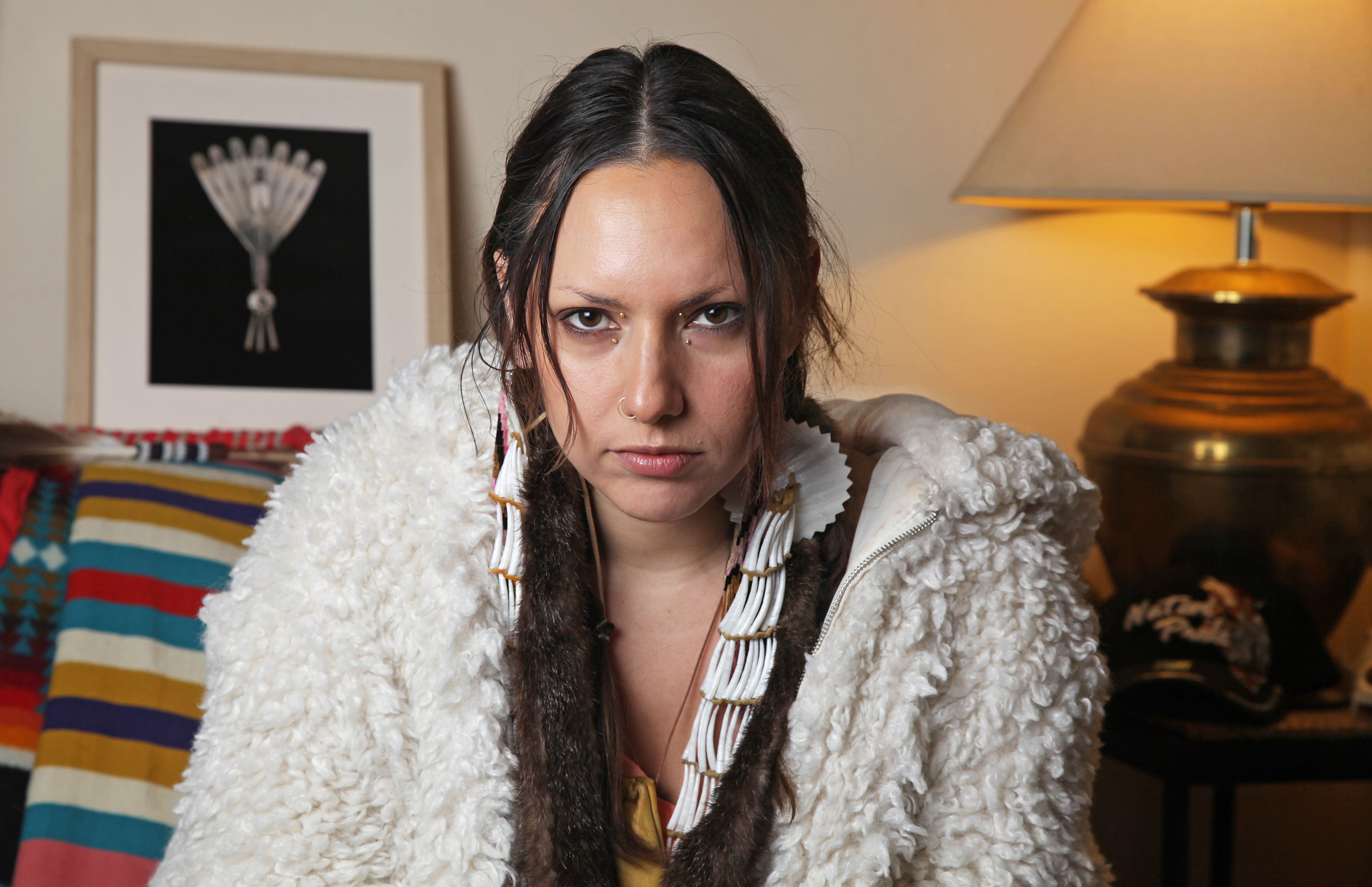 Kite, a young Oglála Lakȟóta woman, with facial piercings in a fake fur hoodie stares directly into the camera