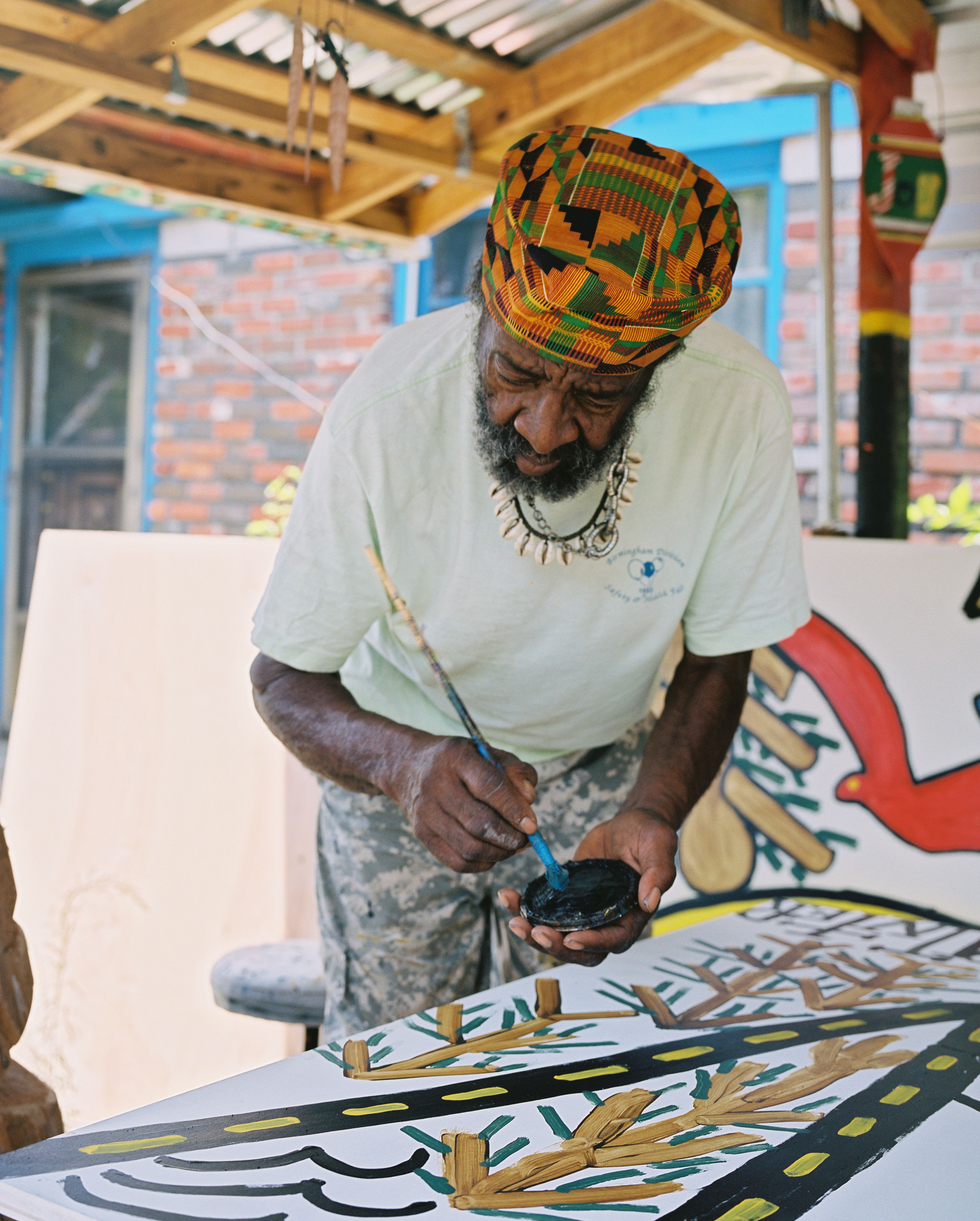 Joe, in a Kente cloth hat and camouflage pants, poised with a brush over a large painting 