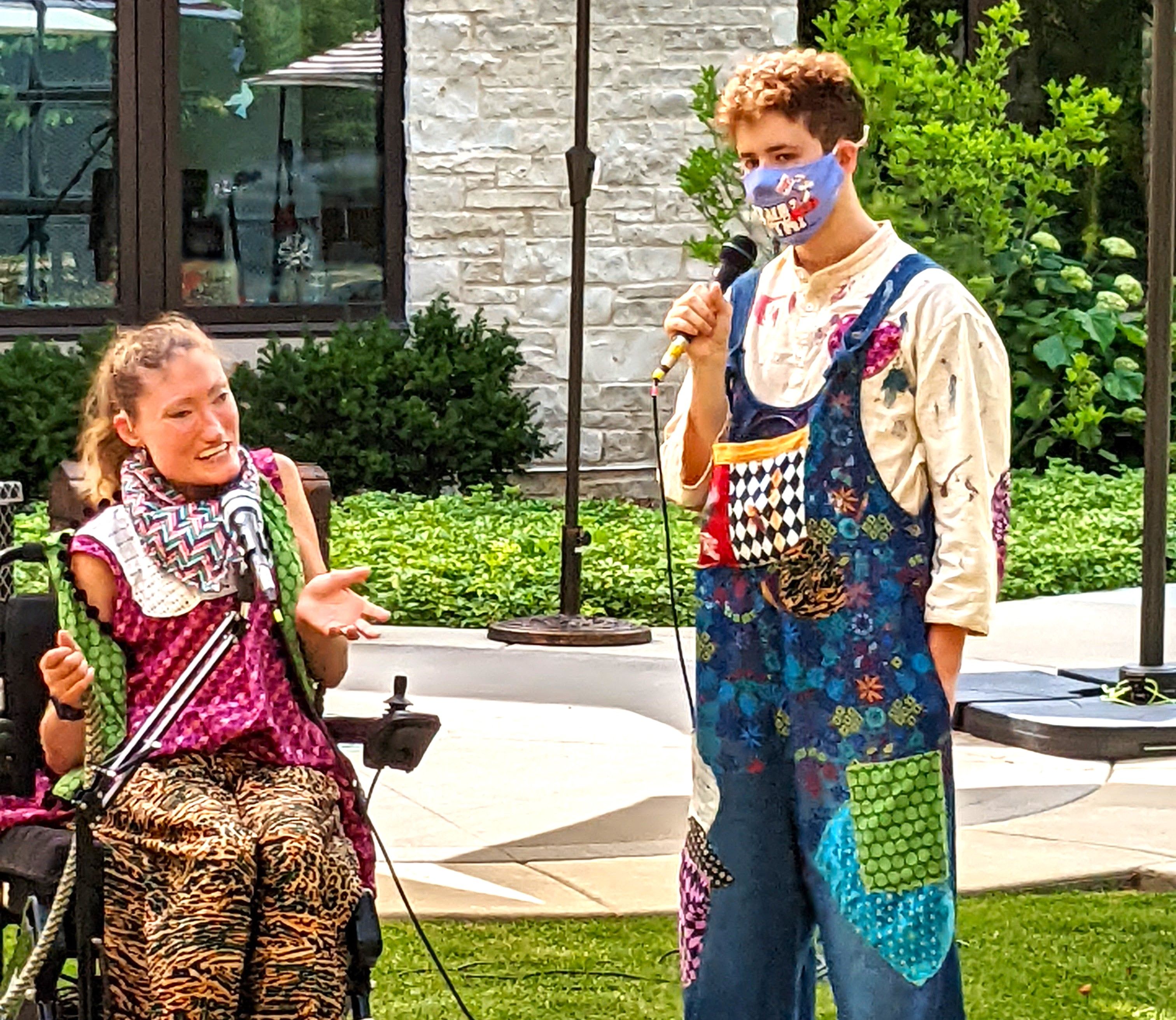 Two performers, one seated in a wheelchair, the other standing with a microphone wearing a facemask and colorful overalls