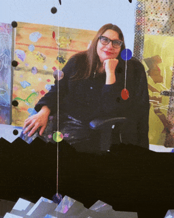portrait of Ranu Mukherjee sitting in a chair in her studio, with artworks leaning against the wall behind her
