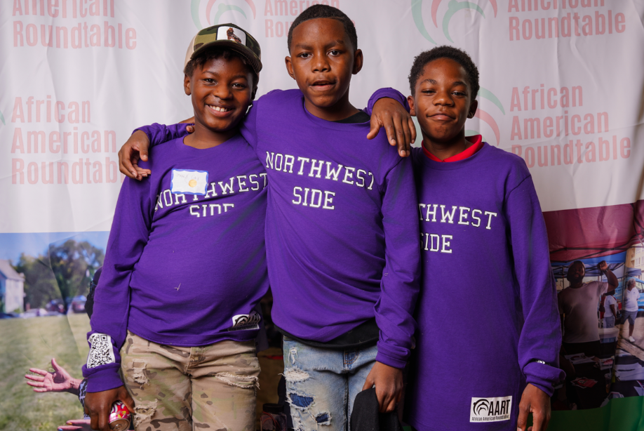 a trio of youth wearing sweatshirts that read "NORTHWEST SIDE" while standing in front of a backdrop printed with a repeat design of the African American Roundtable logo