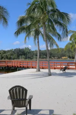 A wooden Adirondack chair on sandy ground facing palm trees and a red footbridge over water.