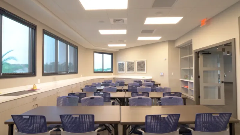 A modern classroom with large windows and blue chairs arranged around tables.