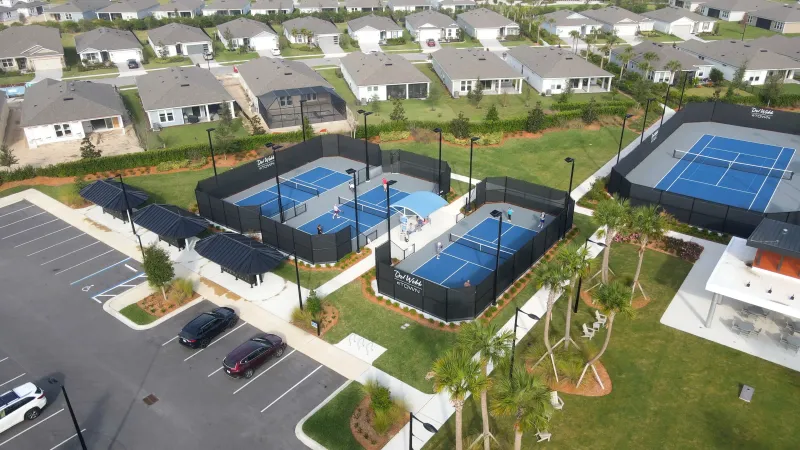 A bird's-eye view of a recreational area with multiple blue sports courts surrounded by high fences, adjacent to a parking lot and residential houses.