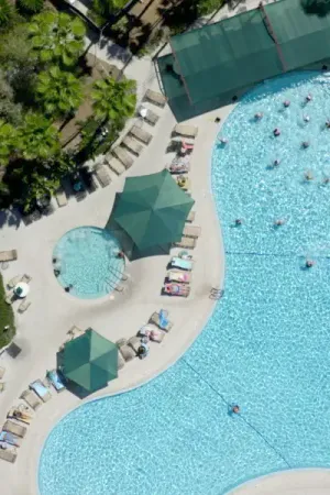 An aerial view of a clear pool, surrounded by people exercising and lounge areas dotted with people relaxing, with teal umbrellas and awnings providing shade and adding a pop of color to the scene.