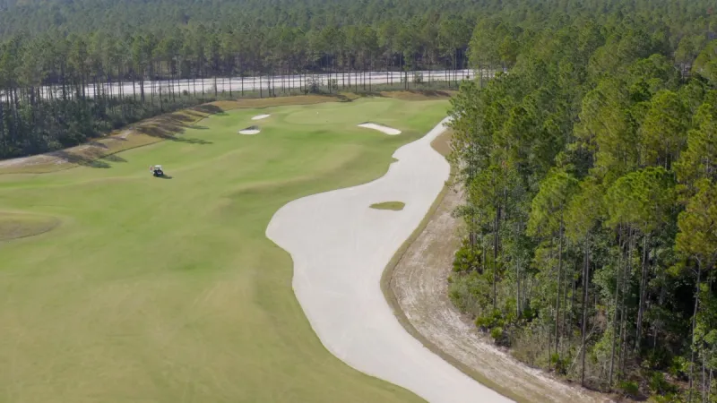 Aerial shot of a curving golf course fairway with sand bunkers, adjacent to a dense forest and a golf cart on the greens.