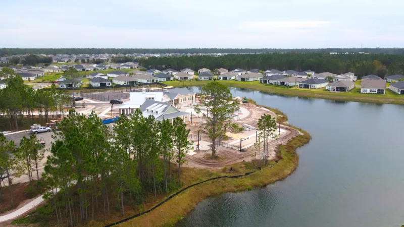 Large green trees in front of the Reverie at TrailMark amenity center set upon a lake with homes in the background.