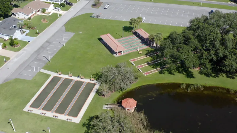 An aerial view of shuffleboard courts, a gazebo by a pond, and surrounding homes with ample green space.
