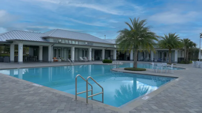 A community pool area with lounge chairs and palm trees, adjacent to a covered patio of a modern clubhouse.