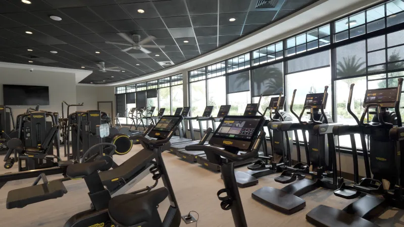 A modern gym with cardio machines, strength equipment, and ceiling fans, well-lit by recessed lighting.
