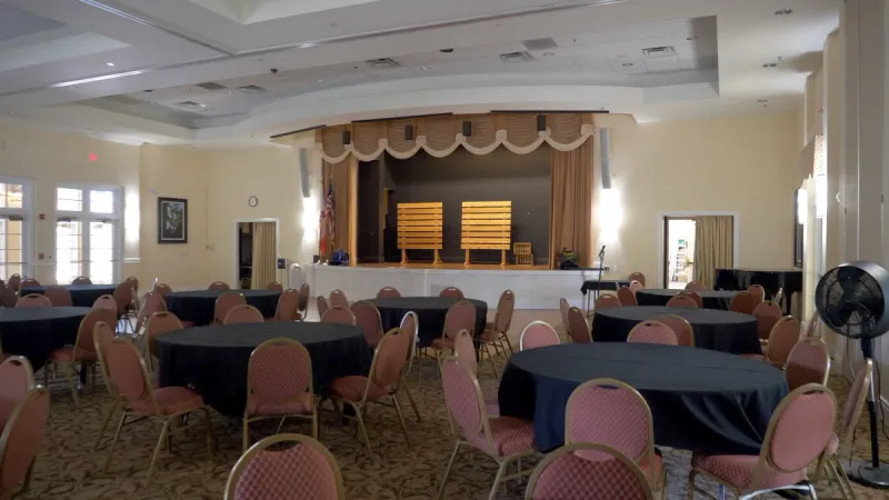 A grand ballroom with round tables and chairs in front of a stage.