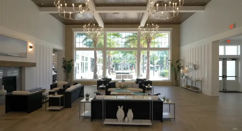 A spacious clubhouse interior with high ceilings, chandeliers, and large windows offering a view of the outdoor seating area.