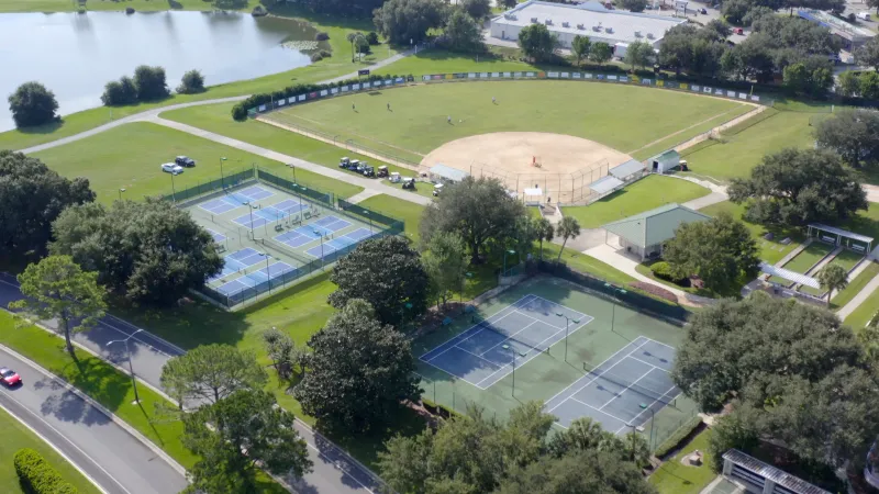 The community sports area with tennis and pickleball courts, a softball diamond, and a pond surrounded by roads and greenery.