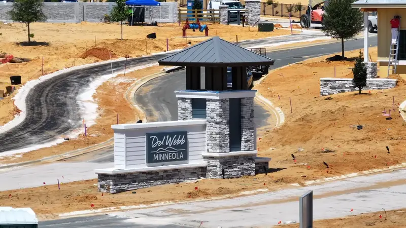 The entrance of Del Webb Minneola with a stylish signpost and a gazebo, set against the construction site with workers actively shaping the community.