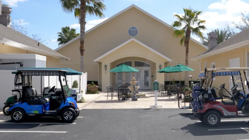 Golf carts parked in front of a community center with tables and chairs providing outdoor sitting.