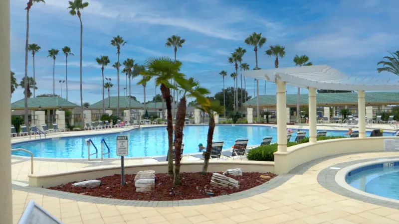 Del Webb Spruce Creek Country Club's pool area featuring ample lounge chairs and tall palm trees in the background.