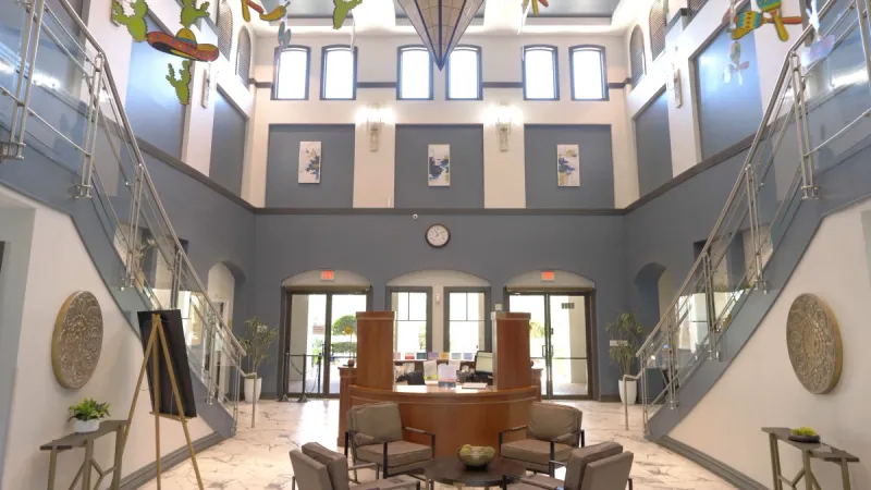 The entrance of a Sun City Center clubhouse featuring an information desk, high ceilings with large windows, and beautifully painted white and gray-blue walls, creating an inviting and spacious atmosphere.