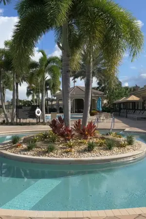 A pool area surrounded by tall palm trees and poolside seating, with a clubhouse in the background under a clear blue sky.