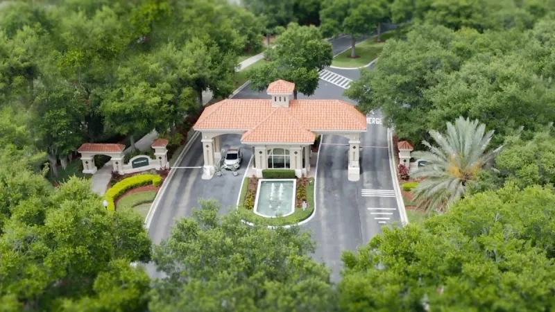 Gated community entrance with a tiled roof over the guardhouse, lush greenery, and a central water fountain.