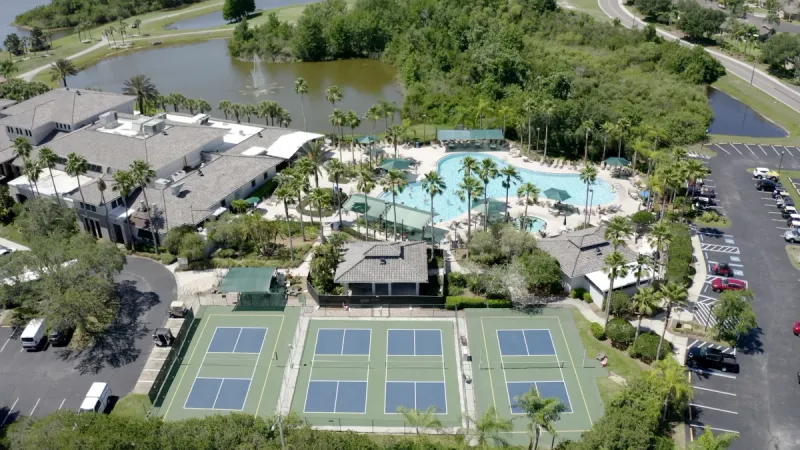 An aerial view of Sun City Center's amenities, showcasing a pool and clubhouse with a parking lot to the side and a water feature in the background.