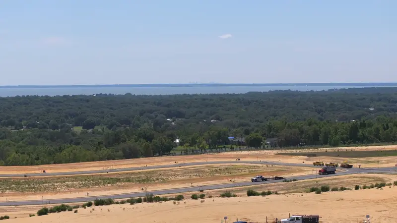 A sweeping aerial view over Del Webb Minneola showing roads curving through the construction site, with a forested landscape and a distant city skyline on the horizon.