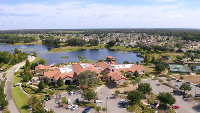 Del Webb Orlando Montecito Clubhouse, a 30,800-square-foot amenity center with parking lot in front, surrounded by lush greenery, sports courts to the side, a lake in the background, and homes in the distance.