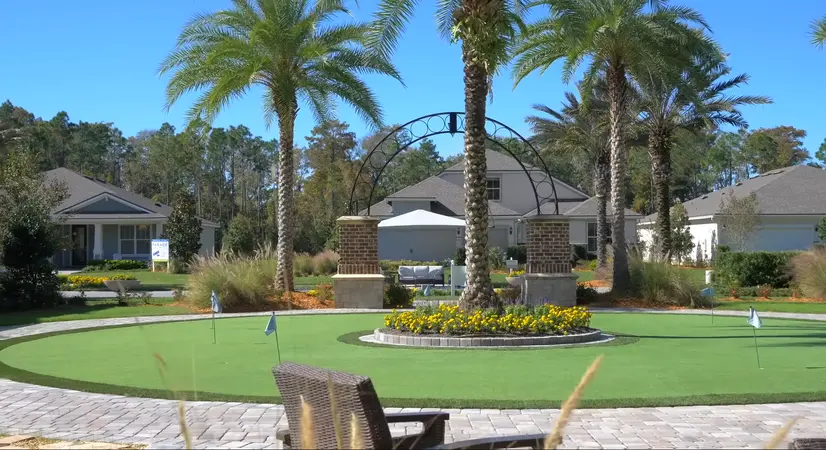 An outdoor putting green in a community space, framed by palm trees and a brick structure with blooming yellow flowers.