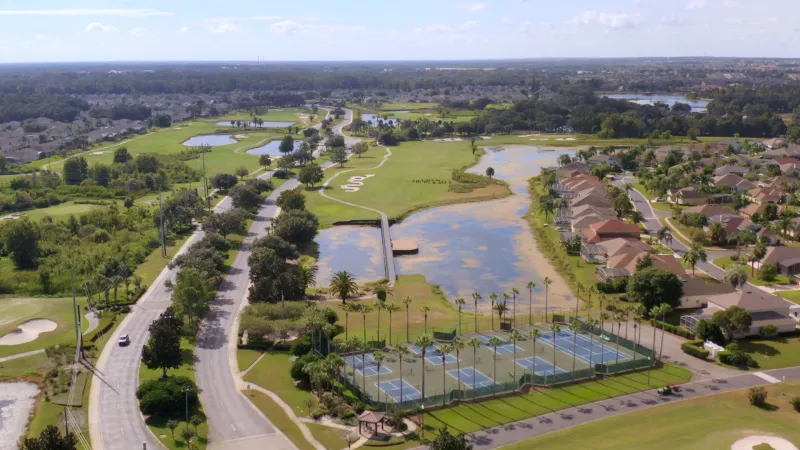 In the foreground, sports courts are surrounded by homes and vibrant landscaping, while the background showcases a scenic golf course.