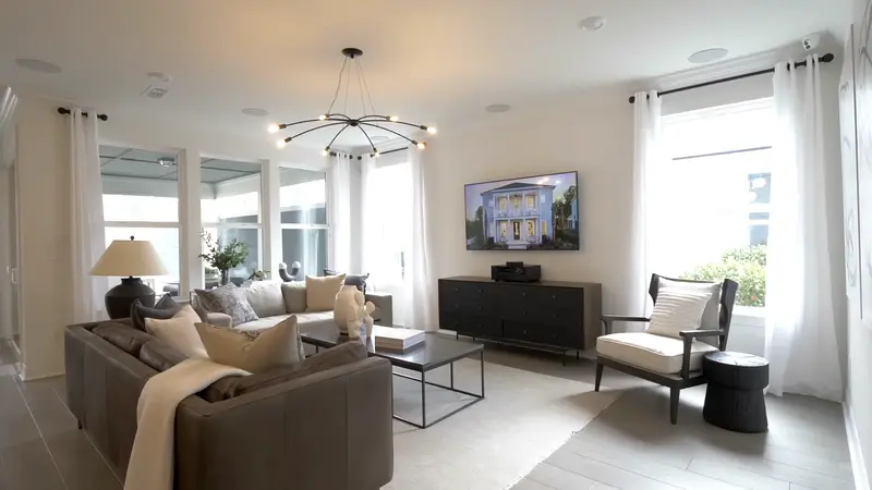 A bright living room with contemporary decor, large windows draped with white curtains, and a TV displaying a home's exterior.