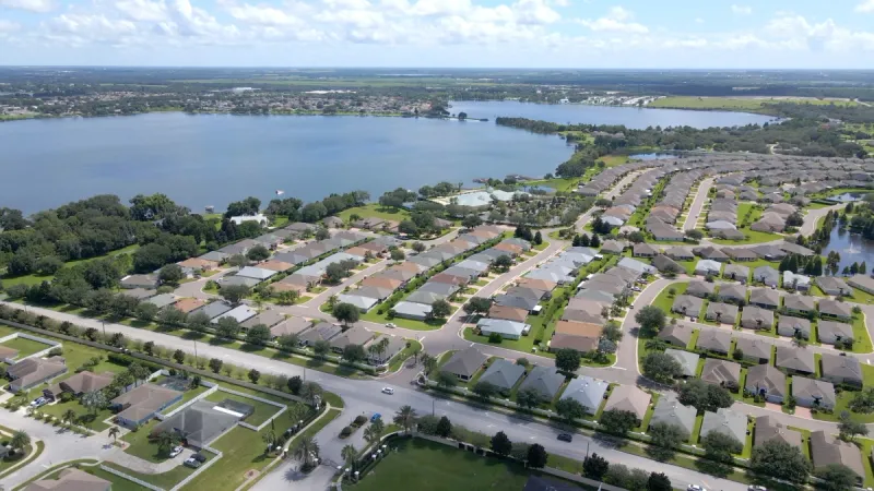 Aerial view of homes situated around a tranquil lake, surrounded by lush green trees in a picturesque neighborhood setting.
