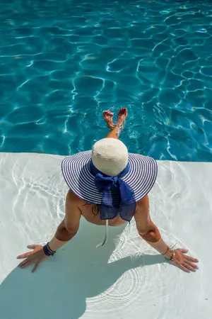 A person in a wide-brimmed hat sits at the edge of a sparkling swimming pool, dipping their feet in the water.
