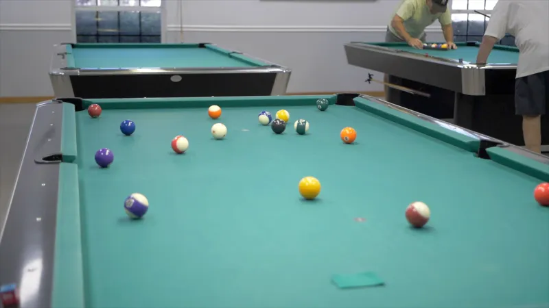 A billiard table with scattered pool balls in the foreground and two individuals playing in the background.