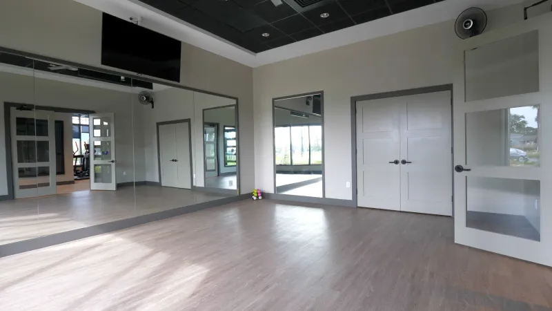 A spacious exercise room at Del Webb eTown with large mirrors, a mounted TV, and glass doors allowing natural light to enter.