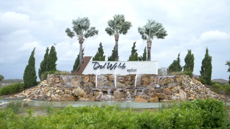 The Del Webb Bexley entrance sign standing atop a rock structure with a cascading waterfall framed by graceful palm trees.