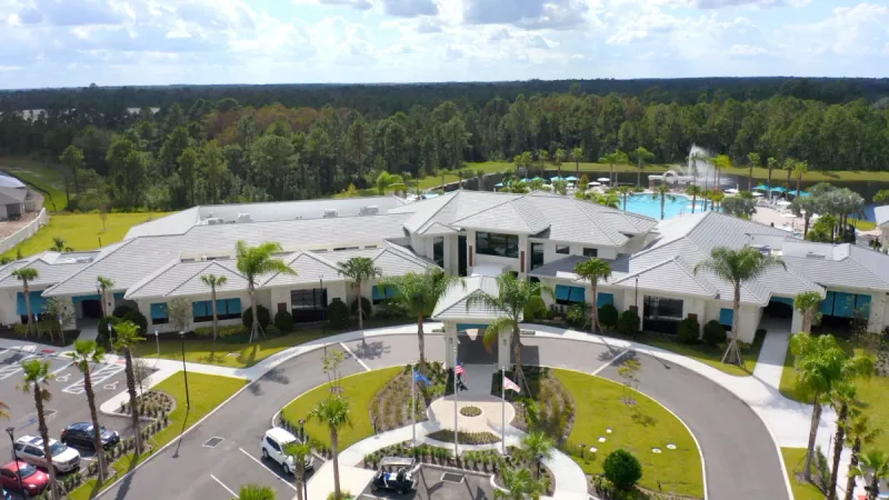 The Del Webb Sunbridge clubhouse featuring a roundabout driveway, palm trees, and well-manicured landscaping, with a pool and trees visible in the background.