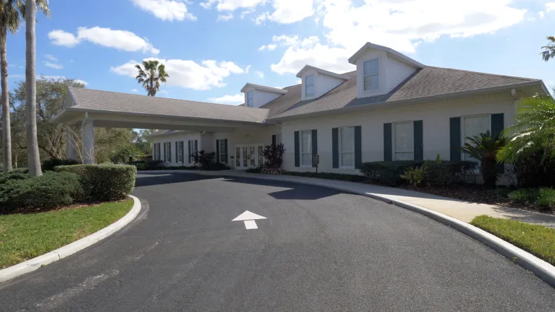 A curving driveway leading up to the Plantation at Leesburg clubhouse, lined with trees and manicured landscaping on either side.