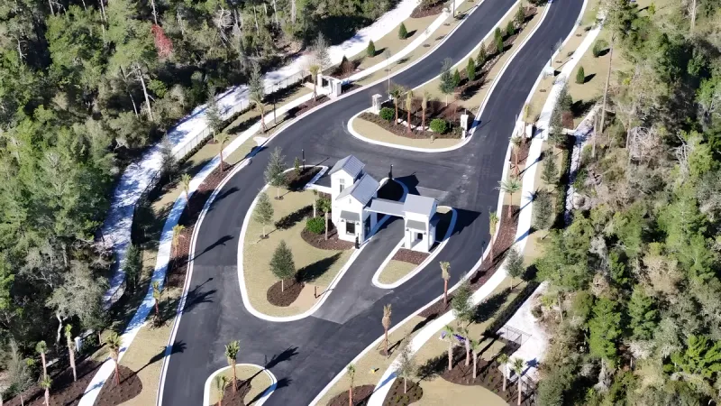 Aerial view of a gated community entrance with palm trees and a winding road amidst lush greenery.