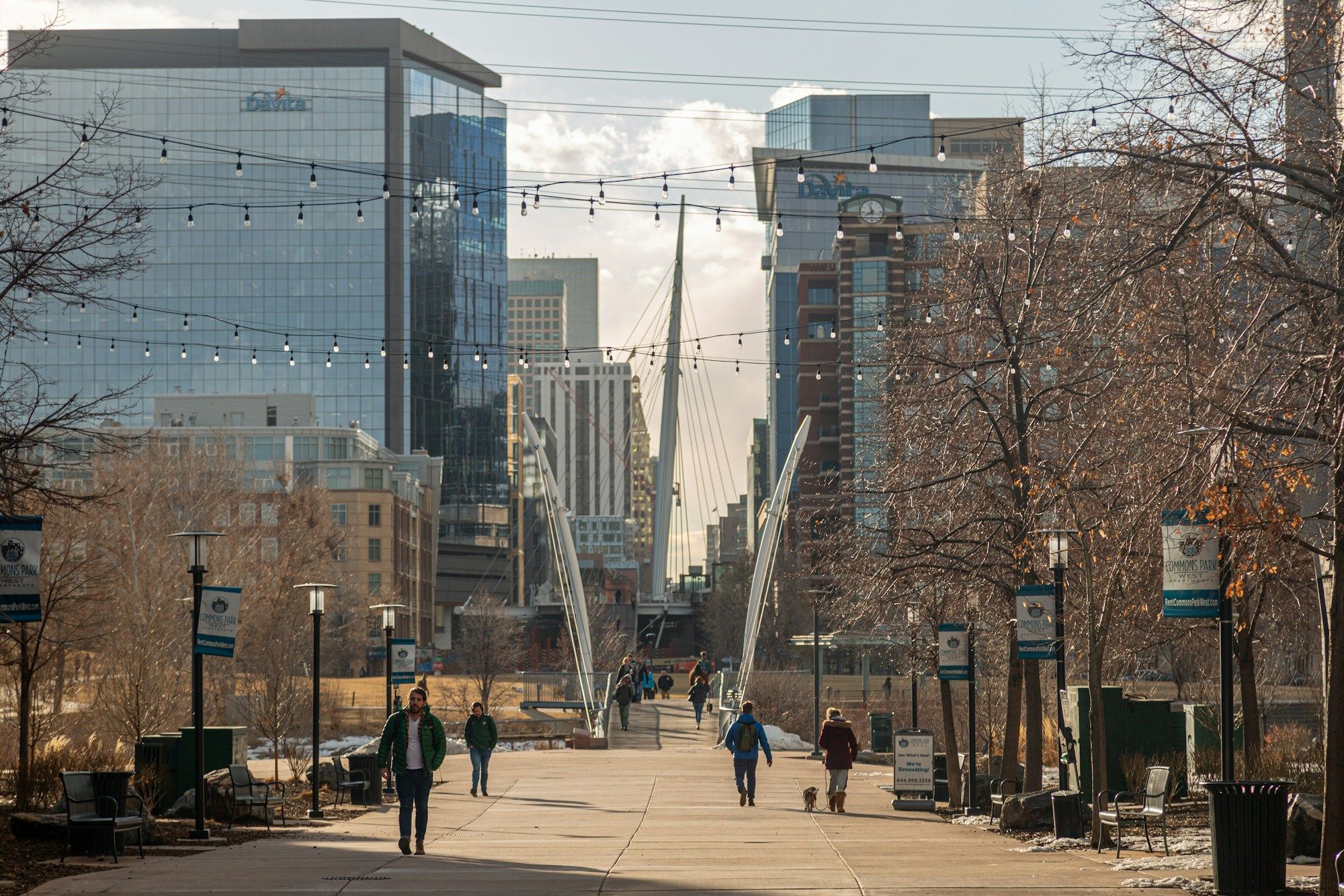 People walking along a pedestrian street in Denver's LoDo under strings of lights, with the white mast of the Millennium Bridge ahead