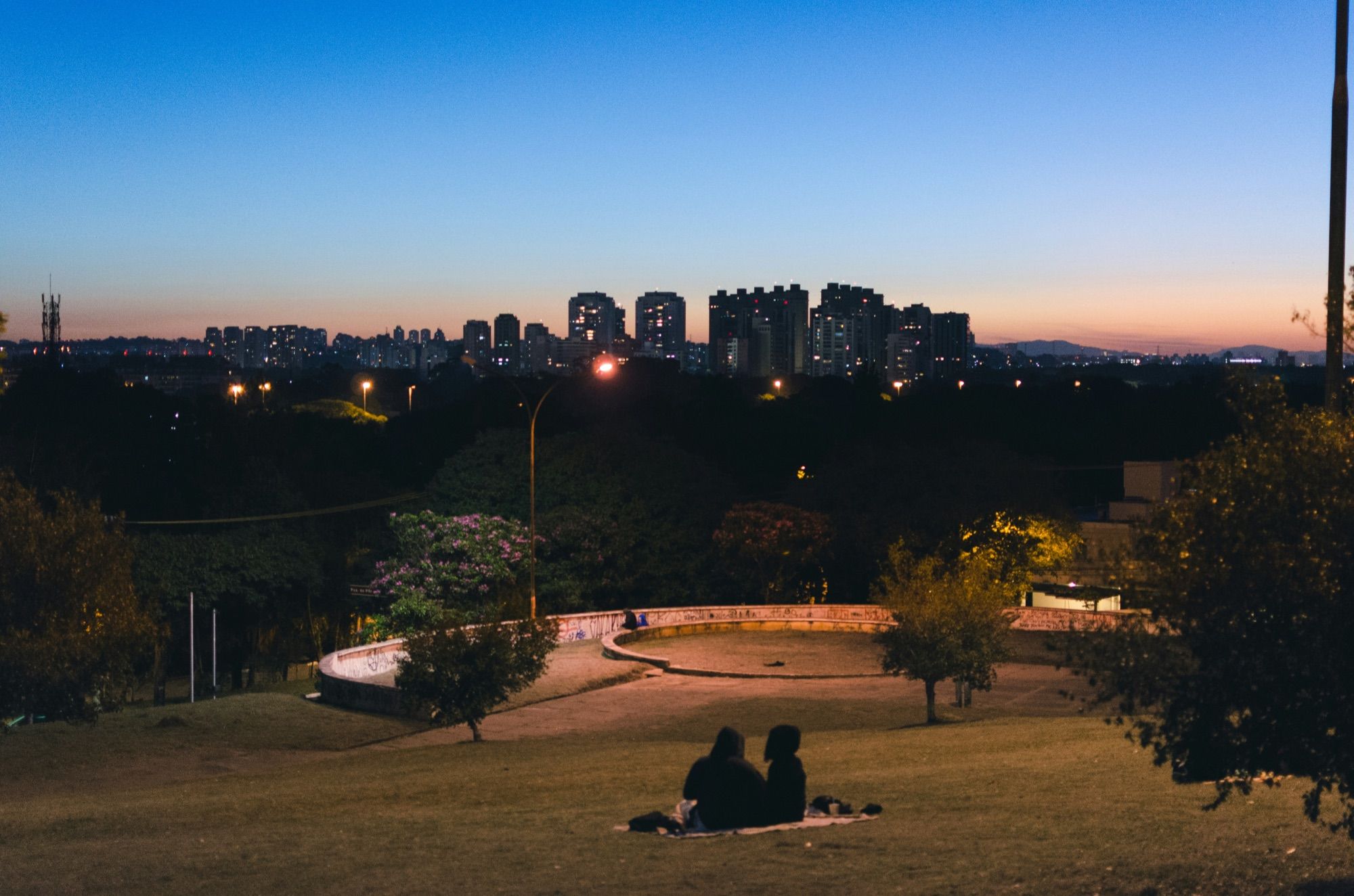 Two people sitting on the lawn of Praça do Pôr do Sol in São Paulo at dusk