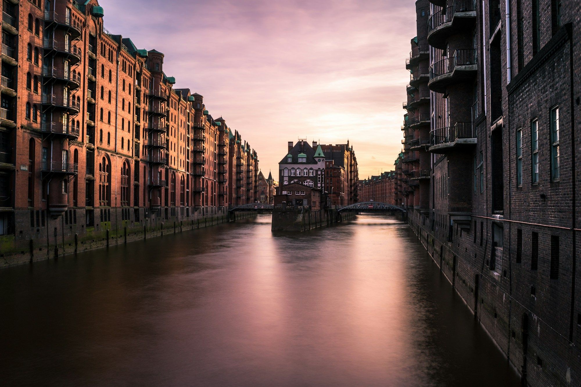 A canal between brick warehouses in Hamburg's Speicherstadt at sunset, with the Wasserschloss at the far end