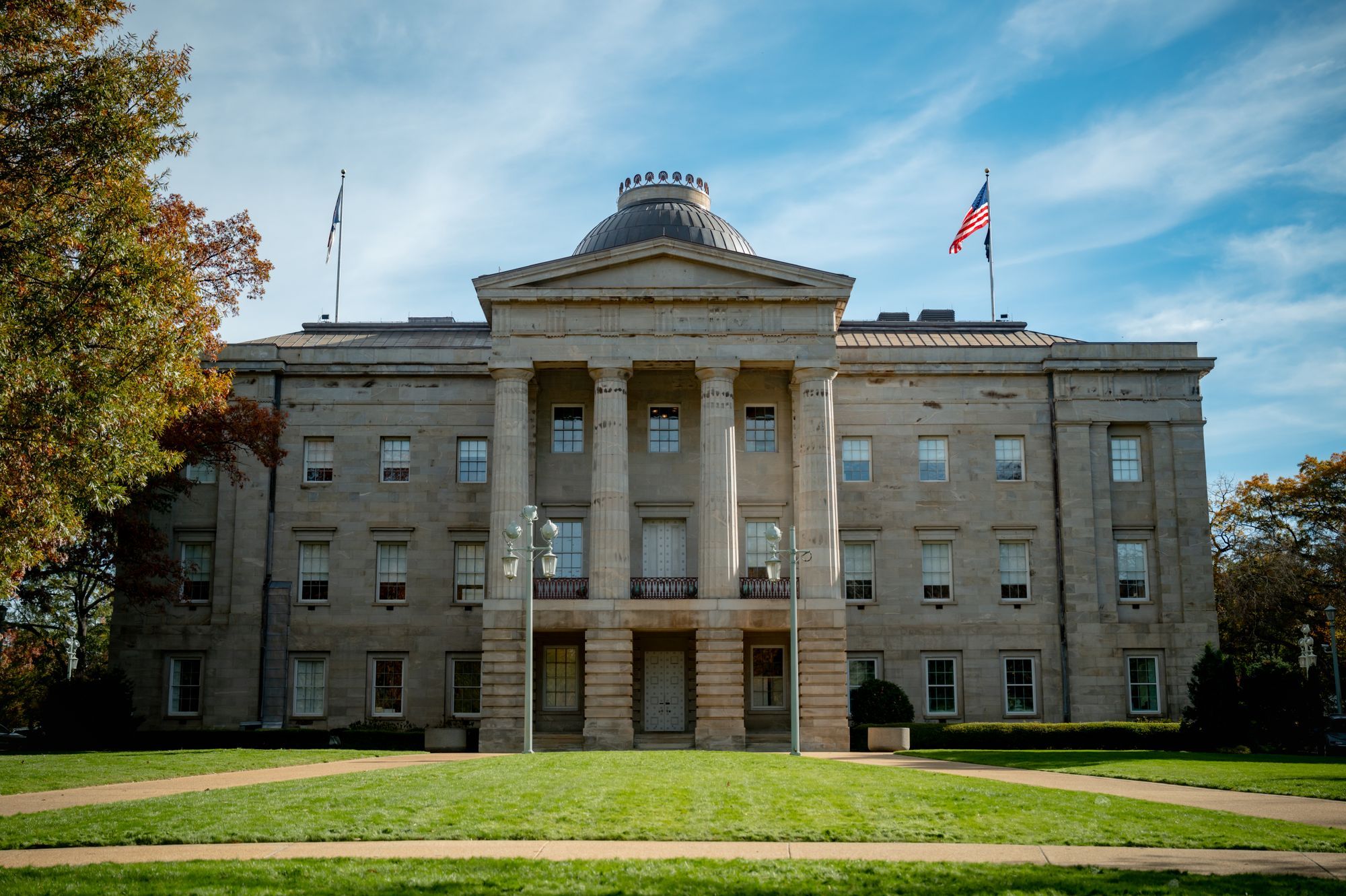 The North Carolina State Capitol in Raleigh, with a lawn in front and autumn leaves at the left.