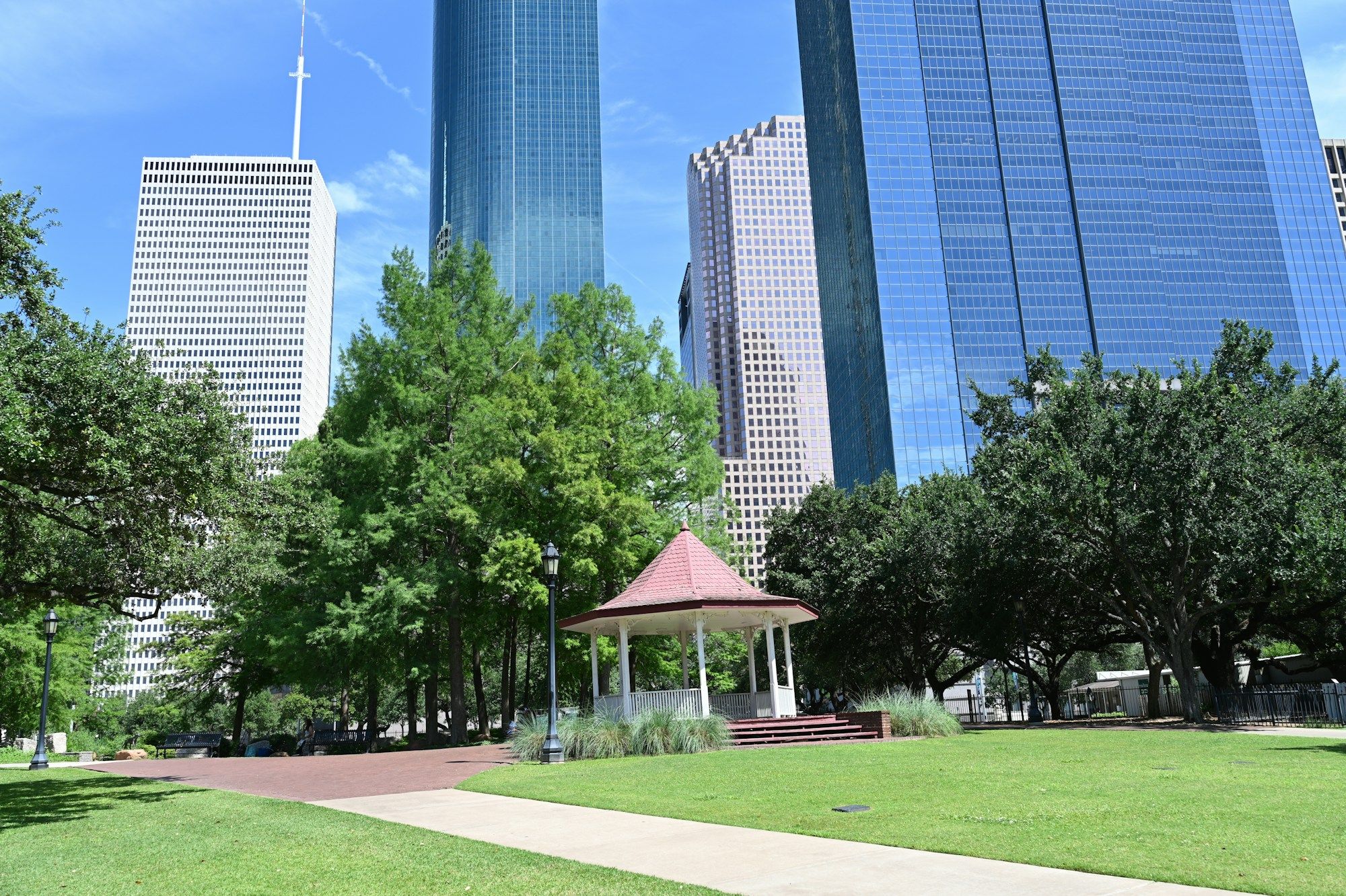 A red-roofed gazebo among oak trees in Sam Houston Park, with the glass towers of downtown Houston behind it