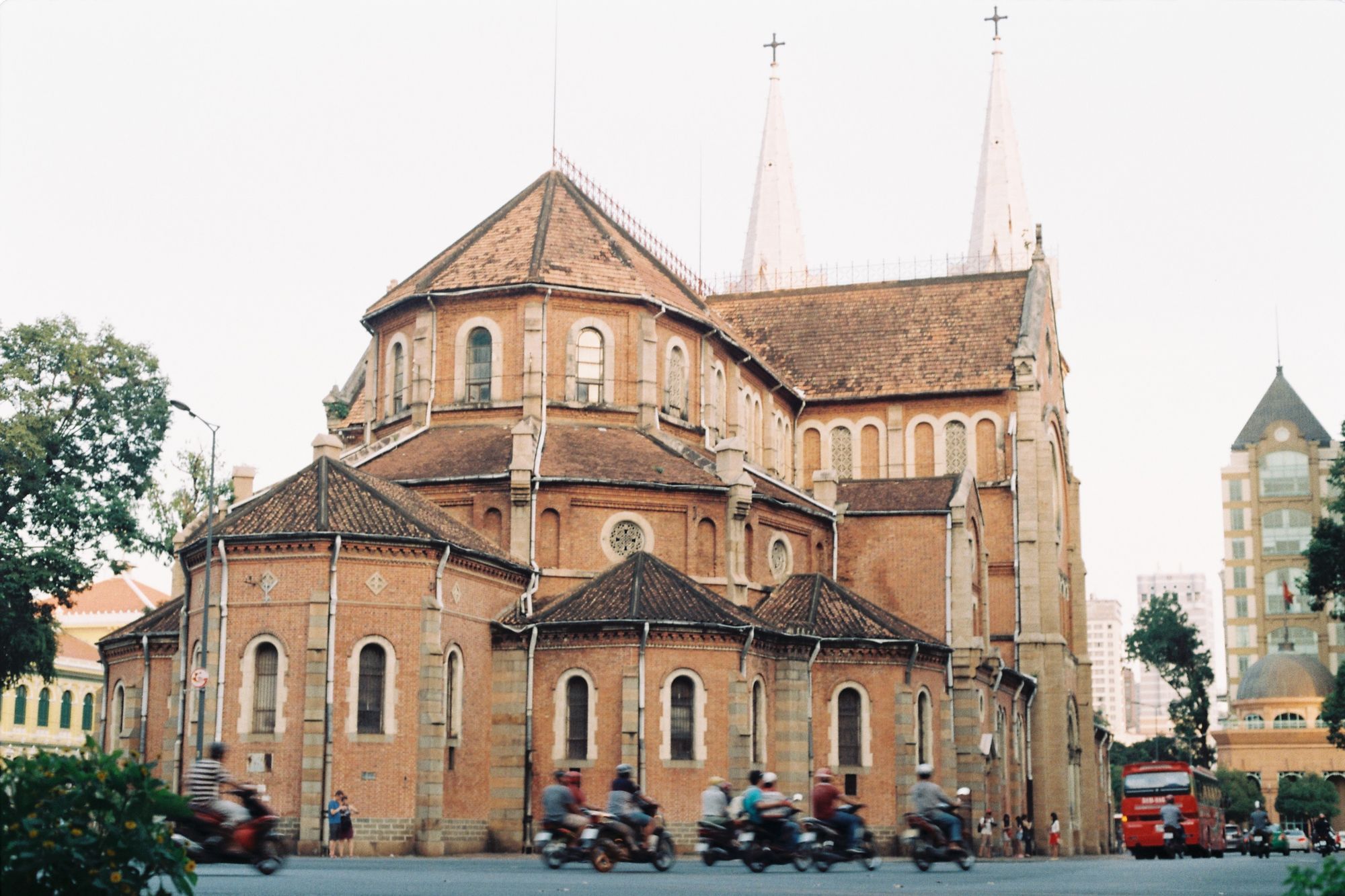 Notre-Dame Cathedral Basilica in Ho Chi Minh City, seen from the side, with brick walls, two spires and motorbikes passing on the street.