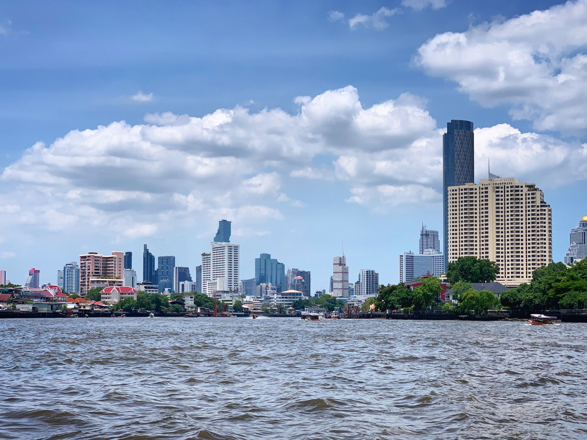 The Bangkok skyline seen across the Chao Phraya River under a partly cloudy sky.