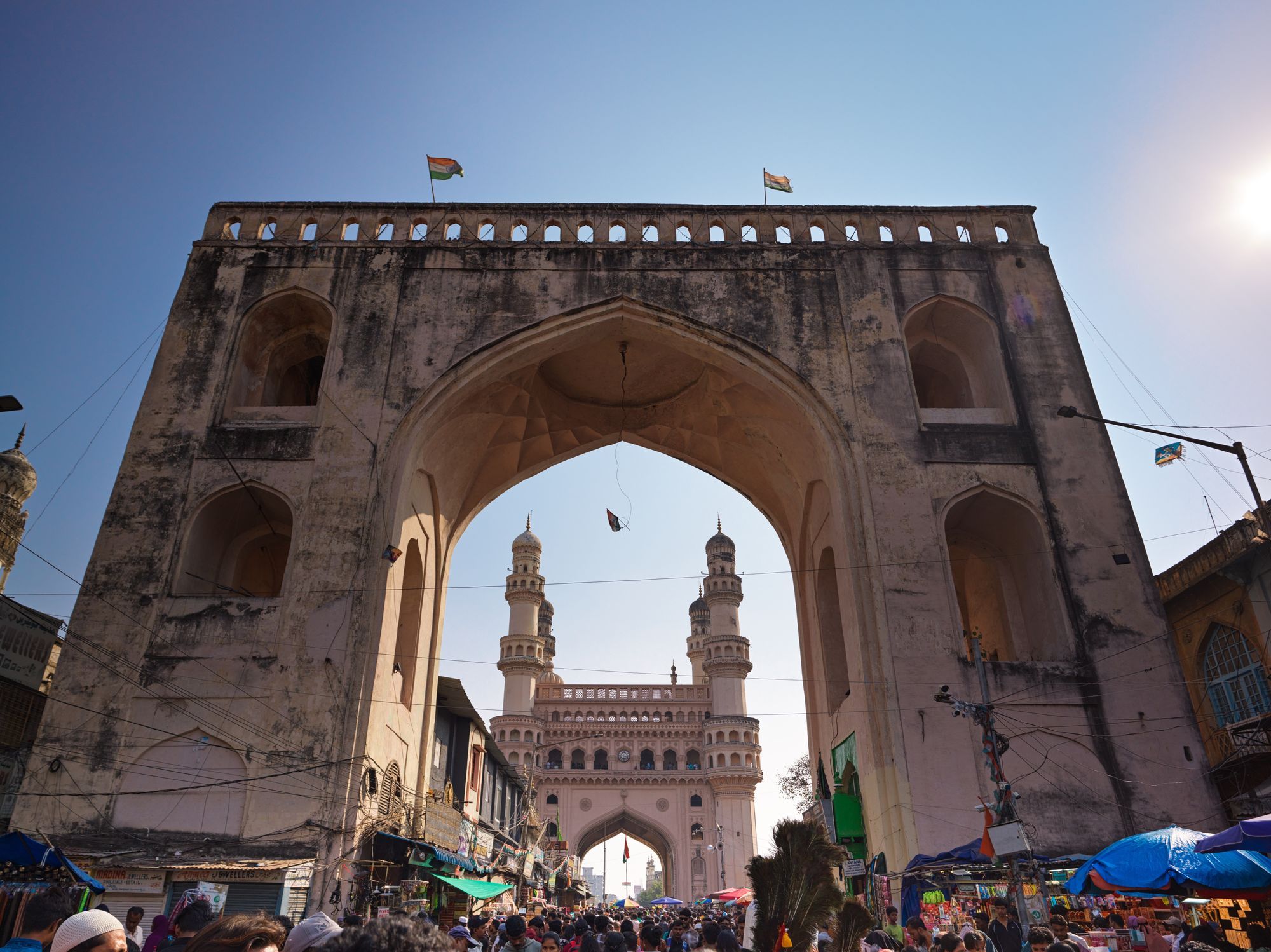The Charminar seen through the Char Kaman gate in Hyderabad's Old City, with market stalls and a crowd below.