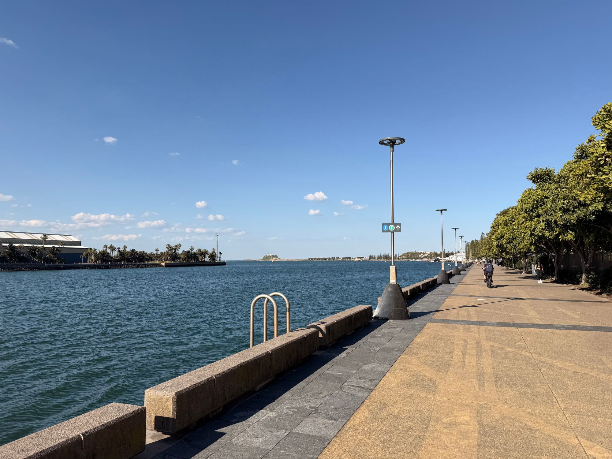 The waterfront promenade in Newcastle running beside the harbor on a sunny day, with the sea on the left and trees on the right.