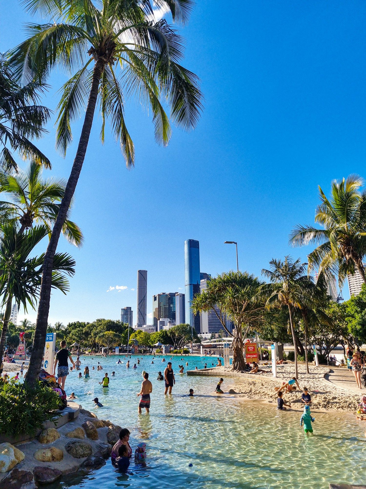 Swimmers in the sandy lagoon of Streets Beach at South Bank in Brisbane, with palm trees and the city towers behind.
